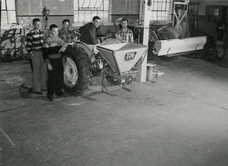 Four students study a piece of farm equipment, a tractor equipped with a seed spreader.