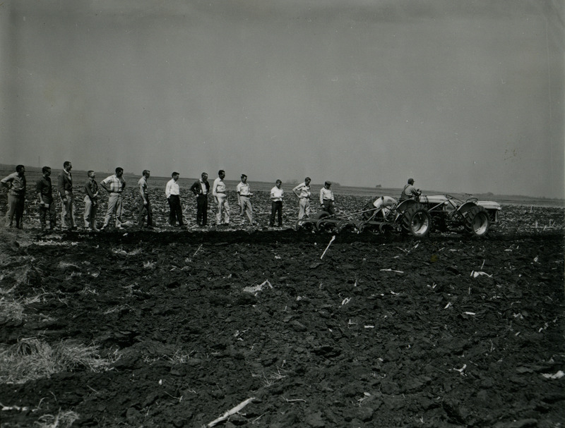 Thirteen students observe someone driving a tractor and cultivating a field.