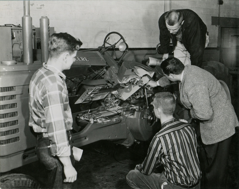 A professor shows three students the mechanics of a John Deere tractor that has been flipped open to show the inside.