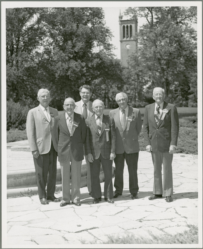 Professor Burnet stands in the back row with five older men wearing ribbons that say "Fiftieth" by the Four Seasons Fountain with the Campanile in the background.