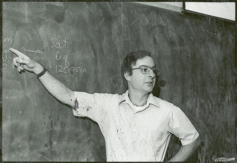 Peter Reilly is standing before a blackboard lecturing to an unseen audience. He is pointing to a formula written on the blackboard.