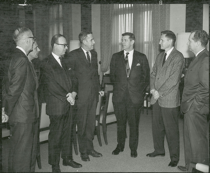 Seven men in suits are engaged in discussion. They are standing in a formal conference room. Iowa State University President, William Robert Parks is standing fourth from the left.