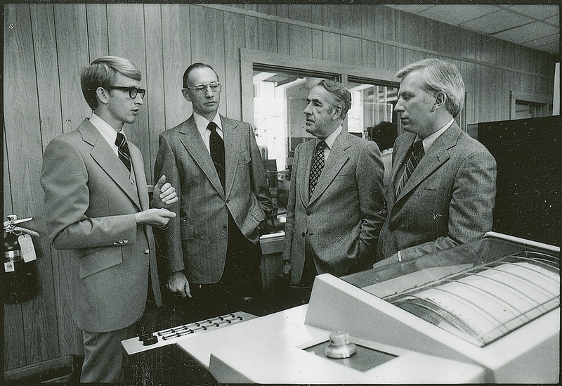 Four individuals are standing in front of the computer. The computer is in the foreground and men are in the background.