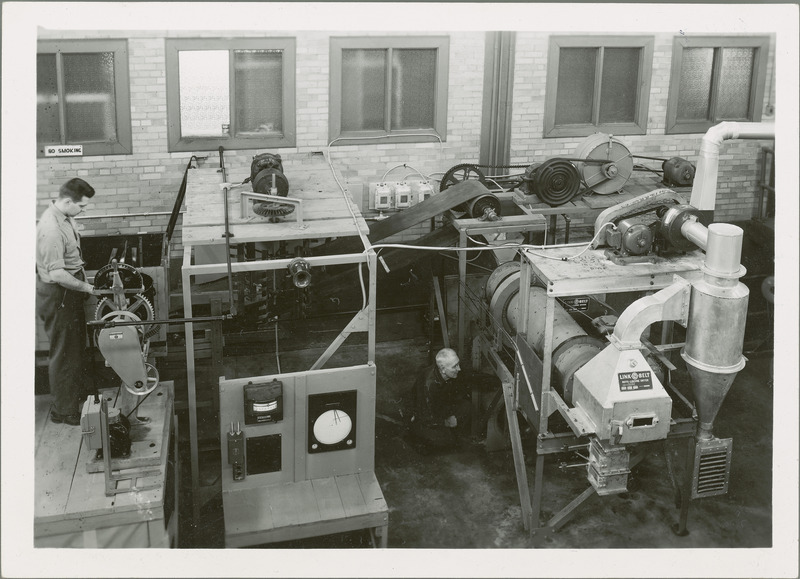 E.C. Kapusta and George West are both working on a large piece of equipments. There are Brown gauges in the foreground and a system of belts and pulleys in the background.