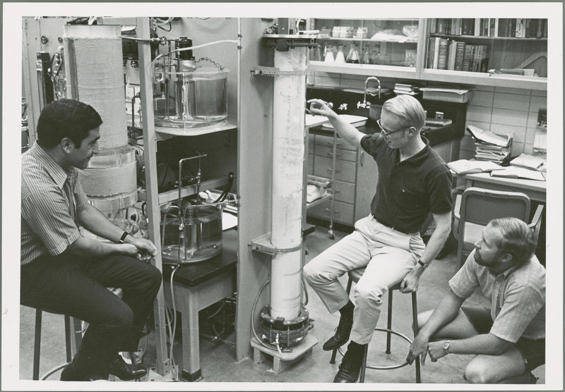 Thomas D. Wheelock and two other researchers are in the chemical engineering lab examining a tall tube shaped apparatus.