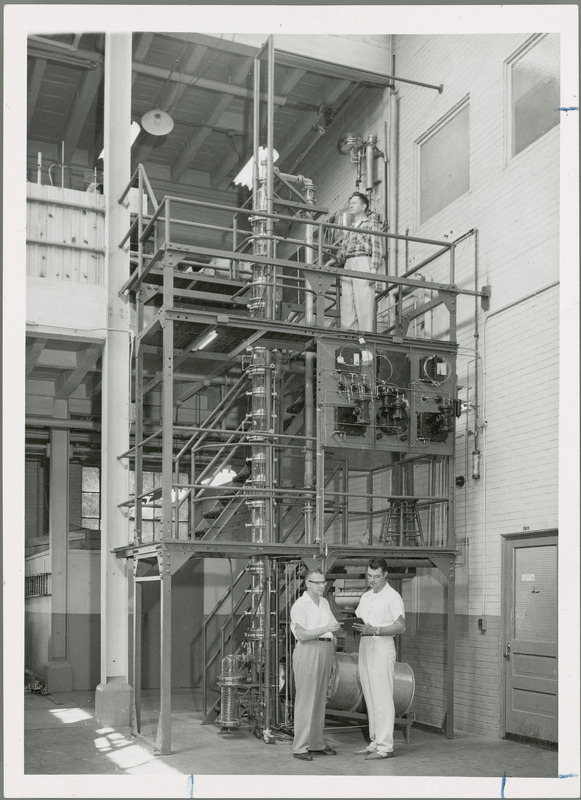 Two researchers are standing before a bubble cap column. The column is surrounded by metal scaffolding and stairs lead to the upper levels. One individual is standing on the highest level near the top of the column.