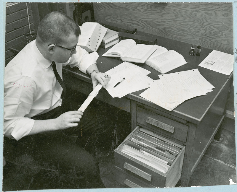 A researcher is sitting at a desk working out calculations on a slide rule. There are papers and several books laying open on desk.