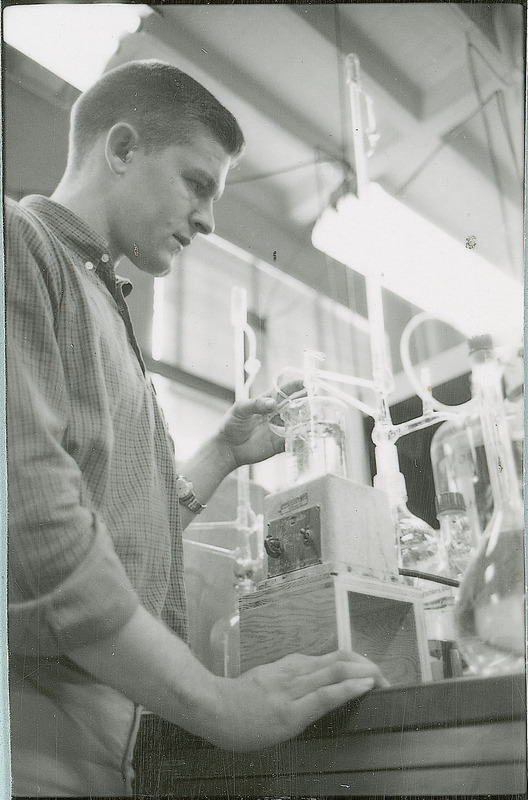 A man is observing a beaker filled with liquid. The beaker is sitting on top of a square control unit. Other tubing and laboratory glassware are connected to the beaker.