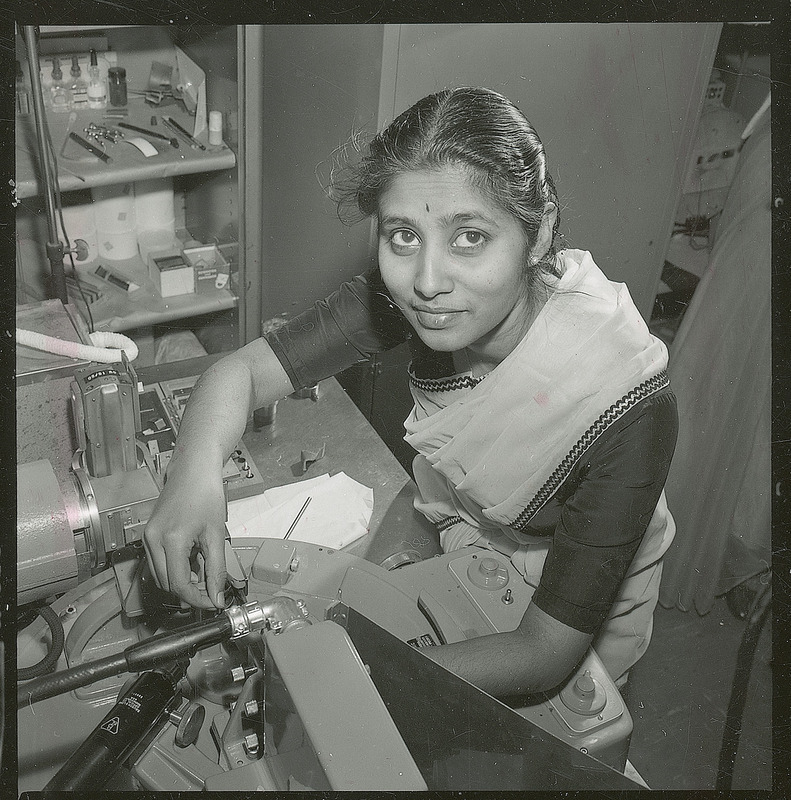 A female graduate student from India is seated at a table working with a piece of equipment in the soils laboratory. At the side of the table is a bookshelf with various laboratory supplies on the shelves. The student is dressed in a tradition sari and wears a bindi mark on her forehead.