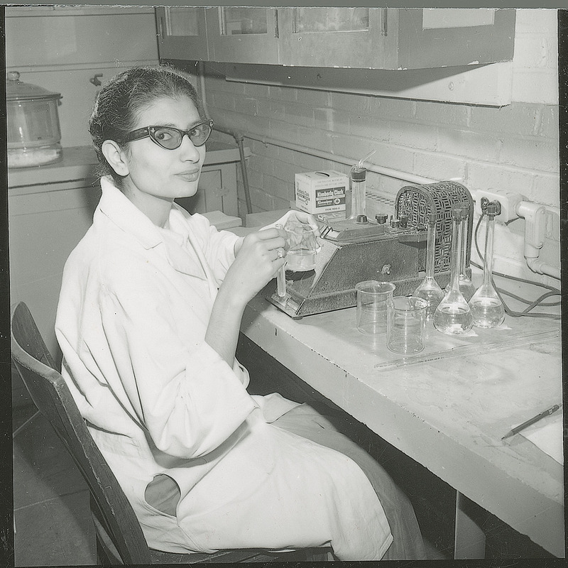 A female graduate student from Indian is seated at the table. She is holding two pieces of glassware. There are additional pieces of laboratory equipments and glassware on the table.