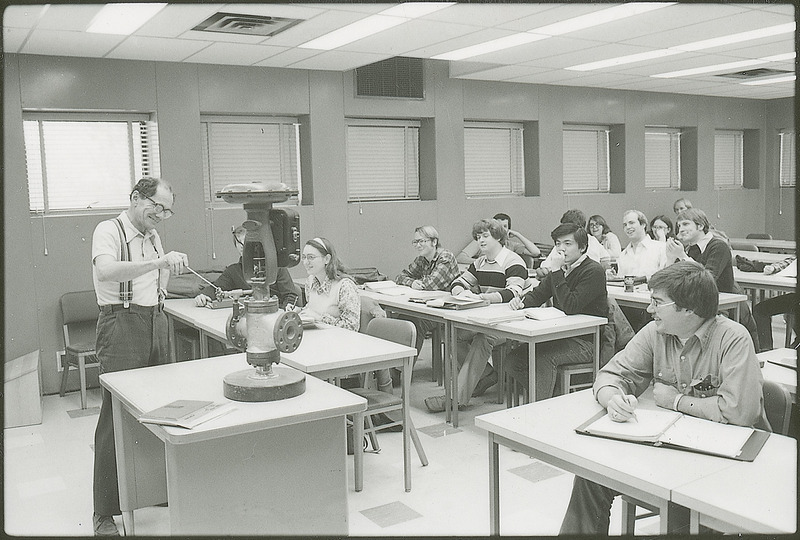 A professor is lecturing to a group of students in a classroom. The subject of the lecture involves a large piece of equipment which is sitting on a table in the front of the room. The teacher is using a pointer to indicate parts of the machine to the students. This photograph was featured in the Chemical Department's descriptive brochure for 1978.