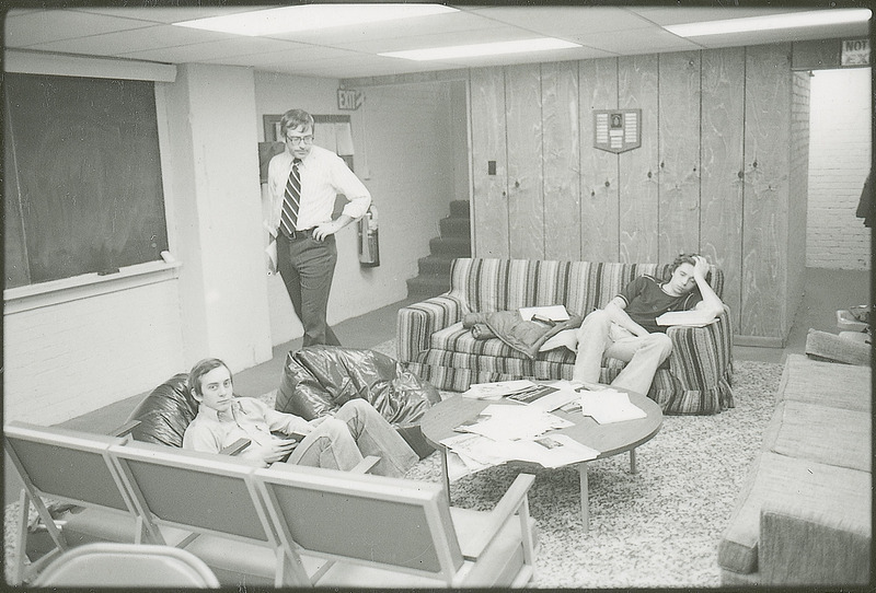 Two students are seated in a lounge type area furnished with a two divans, various types of chairs, a small table and a blackboard on one wall. One student is reading while the other appears to be napping. Another slightly older individual wearing a tie is walking through the area. This photograph was featured in the Chemical Department's descriptive brochure for 1978.