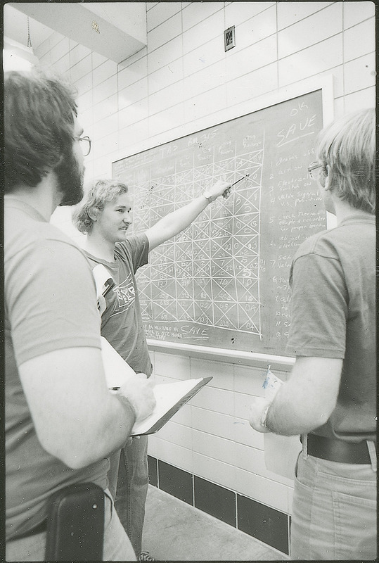 Three students are gathered around a large chart written on a blackboard. One student is pointing to the chart while the other students listen and take notes. This photograph was featured in the Chemical Department's descriptive brochure for 1978.