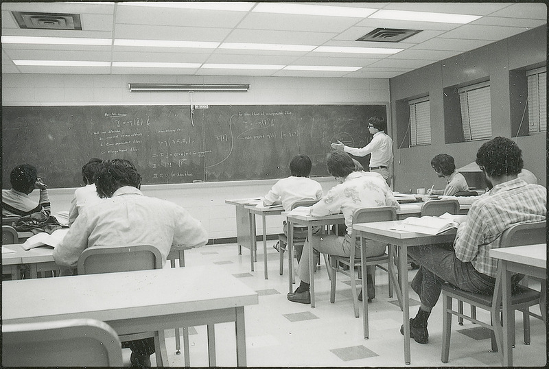 One individual is lecturing to a room of seated students. The lecturer is gesturing to a blackboard filled with notes and various equations. This photograph was featured in the Chemical Department's descriptive brochure for 1978.