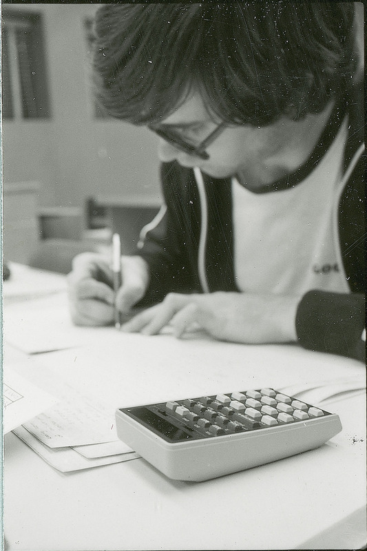 A young man is seated at the table on which there are several sheets of paper and a calculator. The man is writing information on one of the sheets of paper. This photograph was featured in the Chemical Department's descriptive brochure for 1978.