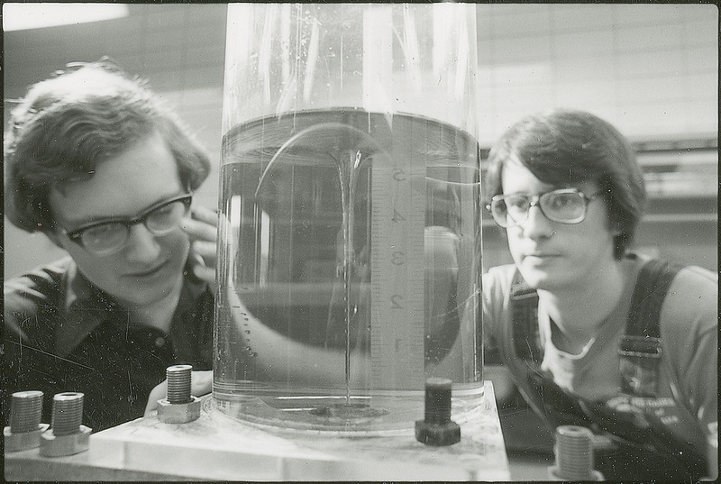 Two researchers are sitting on either side of a glass cylinder filled with liquid. The cylinder is attached to a base and a numeric measuring device is seen through the liquid. This photograph was featured in the Chemical Department's descriptive brochure for 1978.