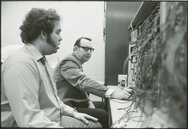 Professor Burkhart and another researcher are seated before a control panel with square gauges. In the fore ground there is a mass of coated wiring.