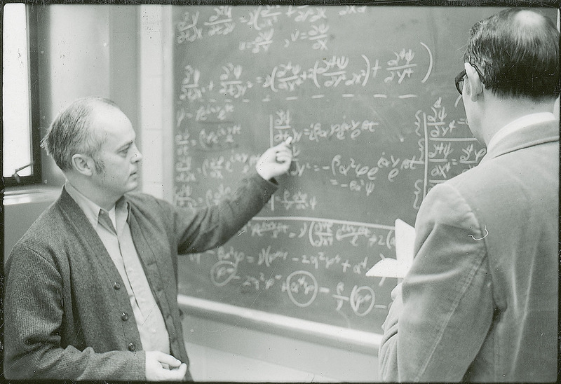 Professors Abraham and Burkhart stand before a blackboard filled with equations. Abraham is using a piece of chalk to point out a specific part of the equation.