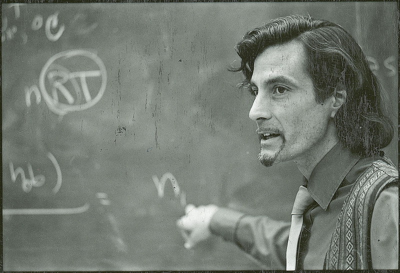 Professor Shuck is standing before a blackboard pointing to a portion of an equation.