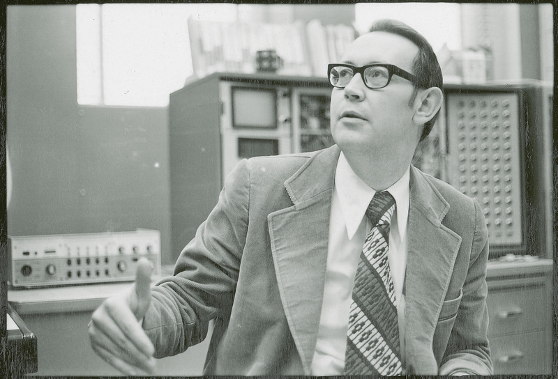 Professor Burkhart is seated in front of two pieces electrical equipment. He is looking upward and gesturing as if in conversation with another individual who is not in the picture,.