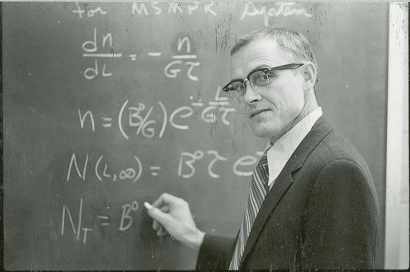 Maurice A. Larson writes out an equation on a blackboard. His gaze is towards the photographer rather than the blackboard.