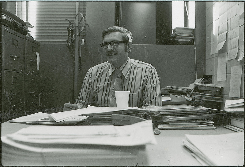 A professor is sitting behind a desk in an office. The desk is covered with various folders, stacks of paper and a Styrofoam cup. There are filing cabinets, a coat rack and windows in the background.