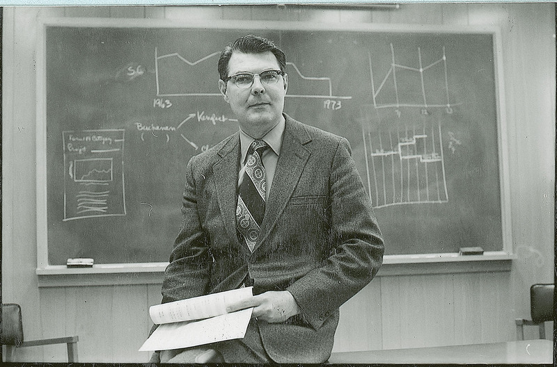 George Burnet is seated on a table before a blackboard covered with various graphics. He is holding several sheets of paper, but is looking directly at the camera.