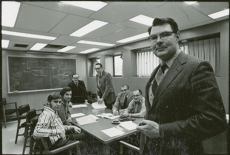 George Burnet other Chemical Engineering professors are gathered around a table. Burnet and two other professors are standing and four professors are seated. Burnet is in the foreground of the image.