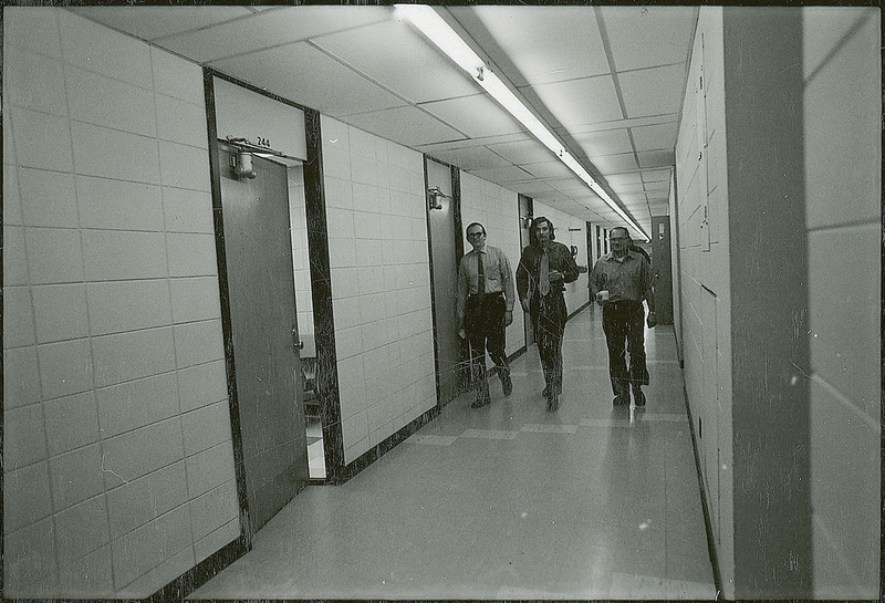 Three professors are walking in a hallway of an academic building.