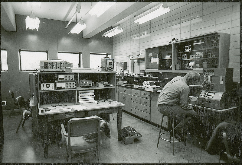 A seated researcher is studying a piece of equipment located before him on a shelf. There are other pieces of equipment on cabinet tops and shelves through the room. An open tool box is laying on the floor near the researcher.