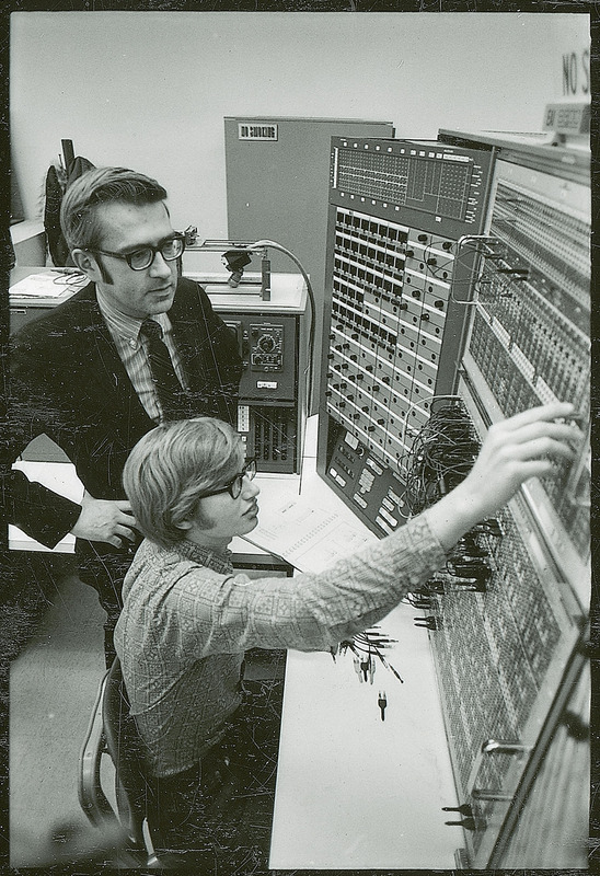 Two researchers are adjusting a control panel for a machine setting on a table. One researcher is standing while the other is seated.