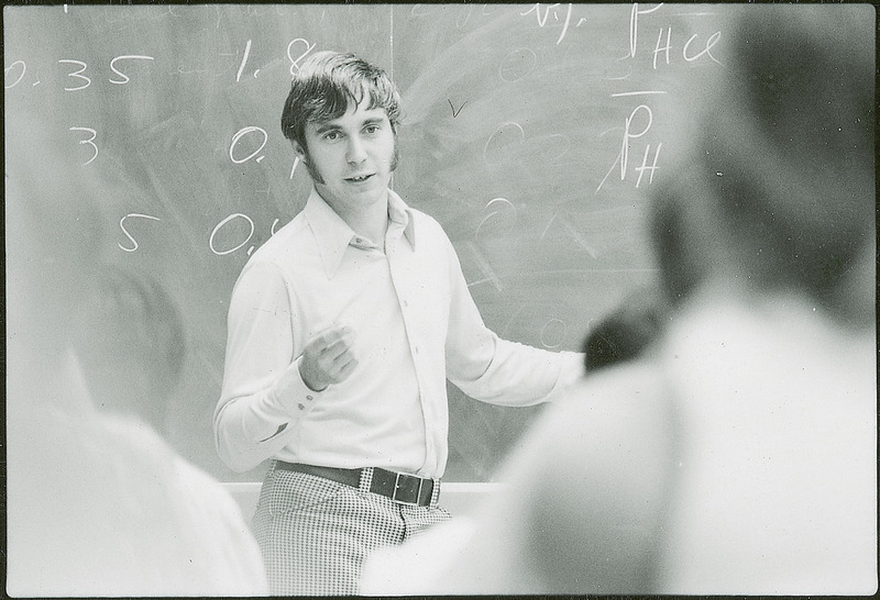 A professor is lecturing to a classroom of students. The professor who is facing the students is in sharp focus, while the students are in soft focus.