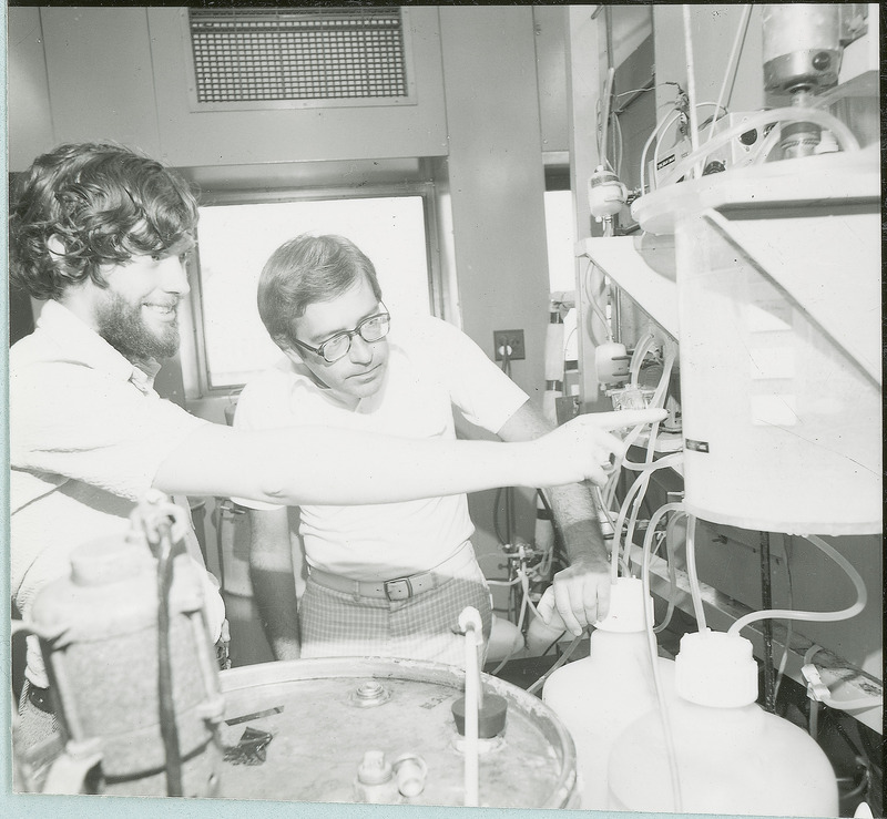 Two researchers stand before a piece of equipment. One of the researchers is pointing to a specific part of the apparatus. This photograph was featured in the Chemical Department's descriptive brochure for 1978.