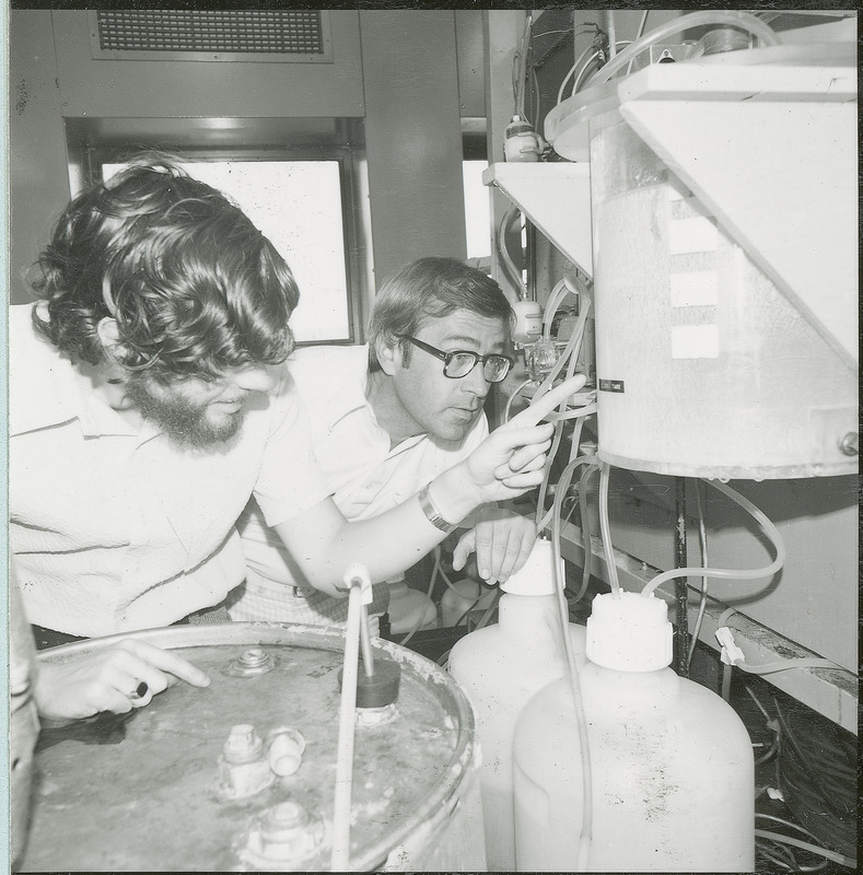 Two researchers stand before a piece of equipment. One of the researchers is pointing to a specific part of the apparatus. This photograph was featured in the Chemical Department's descriptive brochure for 1978.