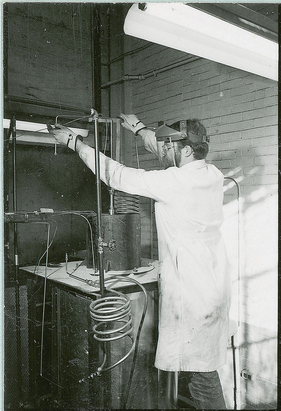 A researcher wearing a full-face shield, heavy gloves and a lab coat works with a piece of equipment that consists of straight and coiled tubing.