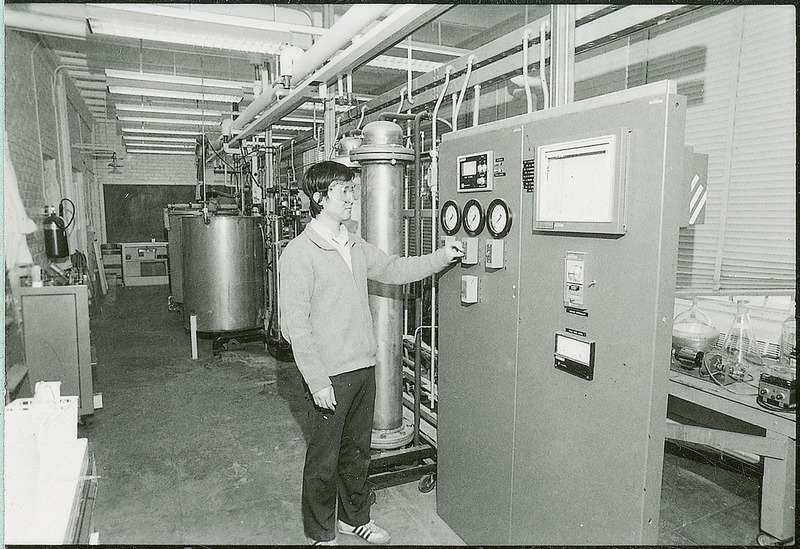 A researcher wearing safety goggles is standing before the control panel of a machine. The control panel has four toggle switches and several dial gauges.