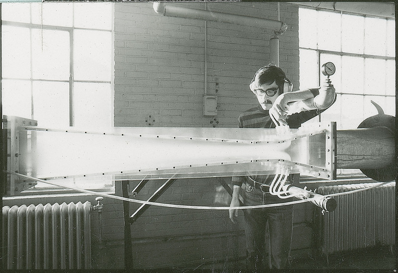 A researcher stands behind a portion of a large piece of equipment. The particular section of the equipment consists of a translucent rectangular shape attached at both ends to large metal pipes.