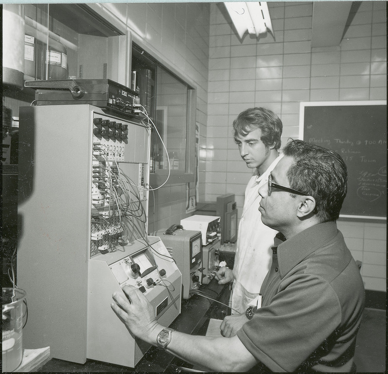 A piece of tabletop equipment is being manipulated by a seated researcher. Another standing researcher is watching. This photograph was featured in the Chemical Engineering Brochure for 1978.