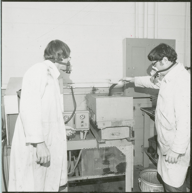 Two researchers stand before a machine labeled, AB roll mill. One researcher is pouring a granular substance into a hopper on the top of the machine. Both researchers are wearing respiration masks. This photograph was featured in the Chemical Engineering Brochure for 1978.