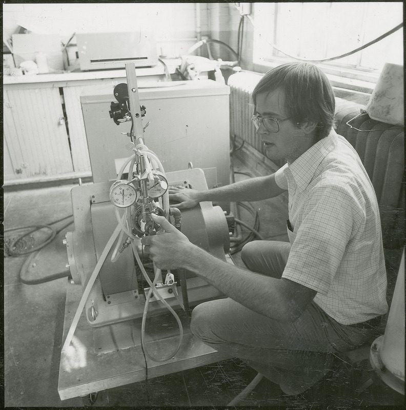 A researcher squats on the floor adjusting a valve while observing a two stage regulator. This photograph was featured in the Chemical Engineering Brochure for 1978.