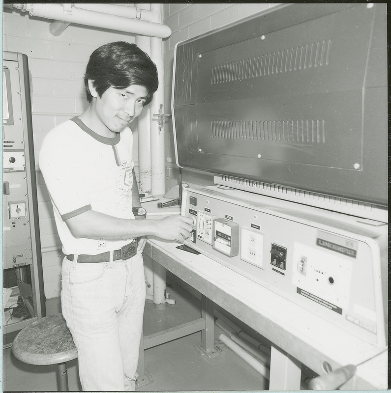 A young researcher is standing before a large piece of equipment. There are various gauges and dials on the bottom half of the apparatus. Other pieces of electronic equipment are in the background. This photograph was featured in the Chemical Engineering Brochure for 1978.