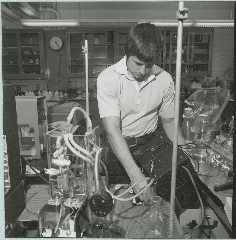 A young researcher is surrounded by various pieces of laboratory equipment. Many of the pieces are attached to each other via tubing. There are glass fronted cabinets in the background. This photograph was featured in the Chemical Engineering Brochure for 1978.