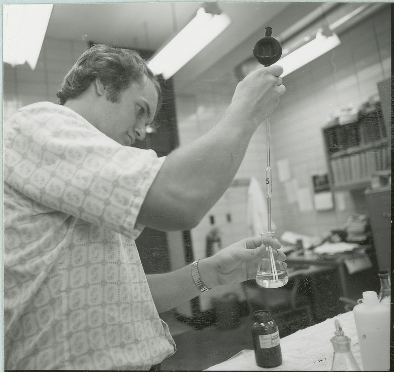 A researcher is transferring liquid between a pipette and an Erlenmeyer flask. Various books and papers are scattered on the table and shelves in the background. This photograph was featured in the Chemical Engineering Brochure for 1978.