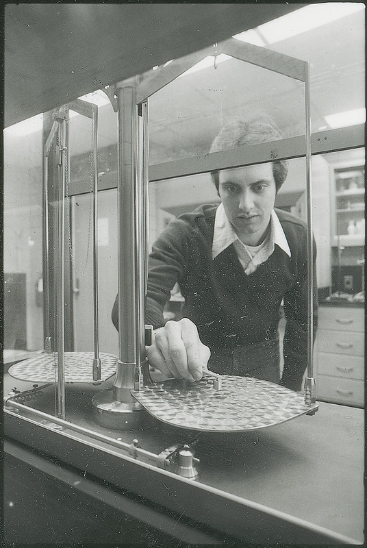 A researcher is using a set of tweezers to place a known mass weight on one pan of a large balance scale. The other pan is empty. This photograph is from the Chemical Engineering Brochure for 1980.