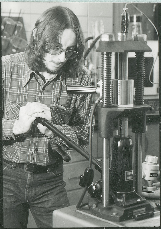 A young researcher is a table top hydraulic press. His hands are resting of the press handle. This photograph is from the Chemical Engineering Brochure for 1980.