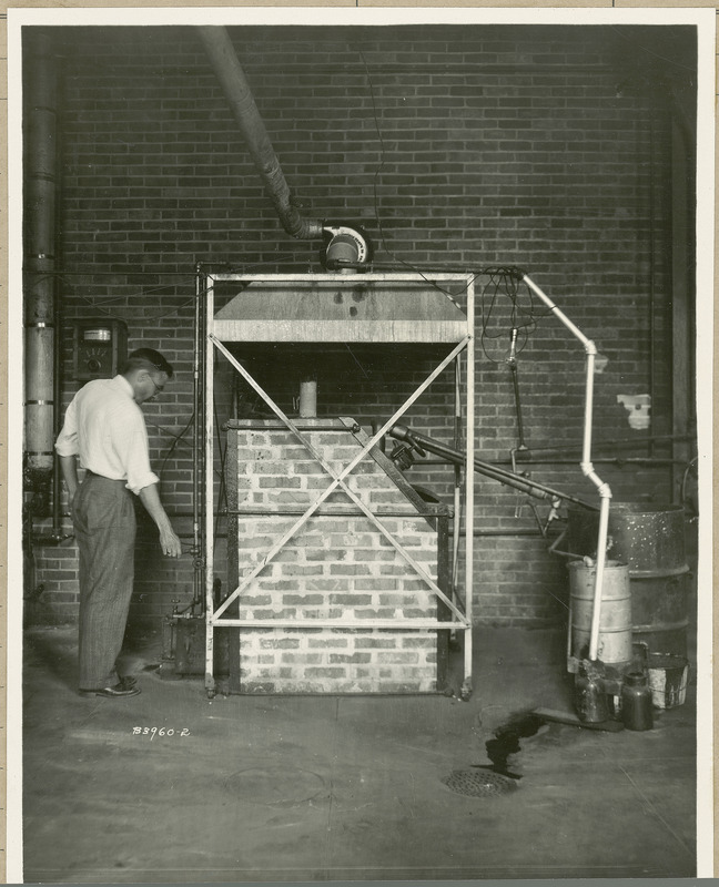 At the center of the photograph is a brick structure enclosed by a metal frame on wheels. The man on the left is operating a switch and there are pipes leading from the structure into two oil drums on the right.