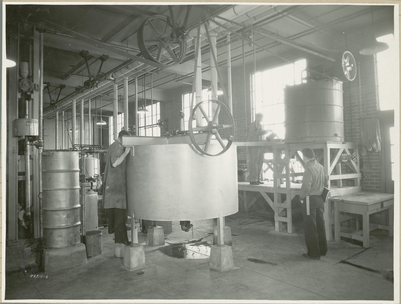 A researcher is looking into a large drum. There is a wheel on top of the drum which is operated by a pulley. A second researcher is pouring a liquid into another machine. A third researcher with a notebook looks on.