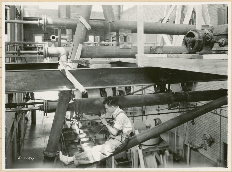 A man is sitting on a pipe while adjusting a valve on other pipe.