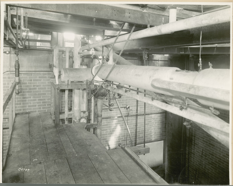 At the center of this view from the balcony level is a large chained pulley. Several long pipes with spigots run horizontally above the wooden floor.