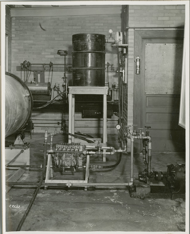 A large drum is partially visible on the left and a large can is sitting on a table in the center. There are a several pipes, valves, and a motor on the lower right. A Sweetland filter is on the floor at the bottom center of the photograph.